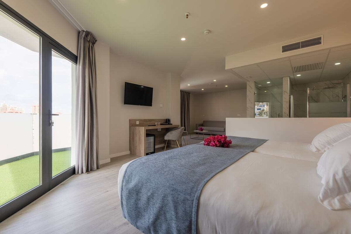 A modern hotel room with a large bed, a desk with a chair, a flat-screen TV, and a balcony with a view of the city.
