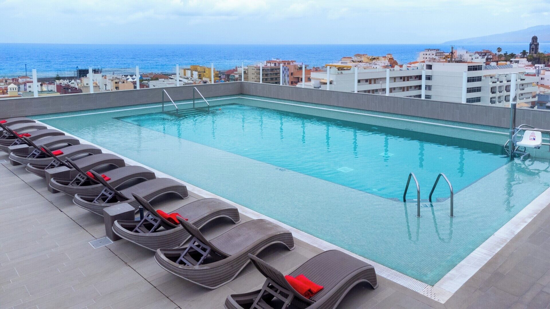A rooftop pool with lounge chairs and a view of the sea and buildings.