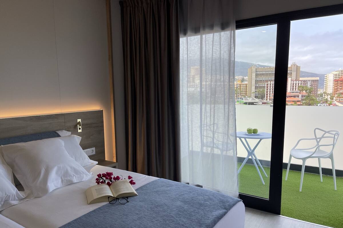 A hotel room with a bed, a small table with two green apples, and a view of a cityscape through large windows.
