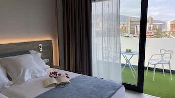 A hotel room with a bed, a small table with two green apples, and a view of a cityscape through large windows.