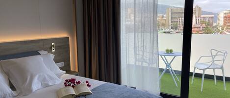 Habitación de hotel con una cama, una pequeña mesa con dos manzanas verdes, y vistas a un paisaje urbano a través de ventanas amplias.