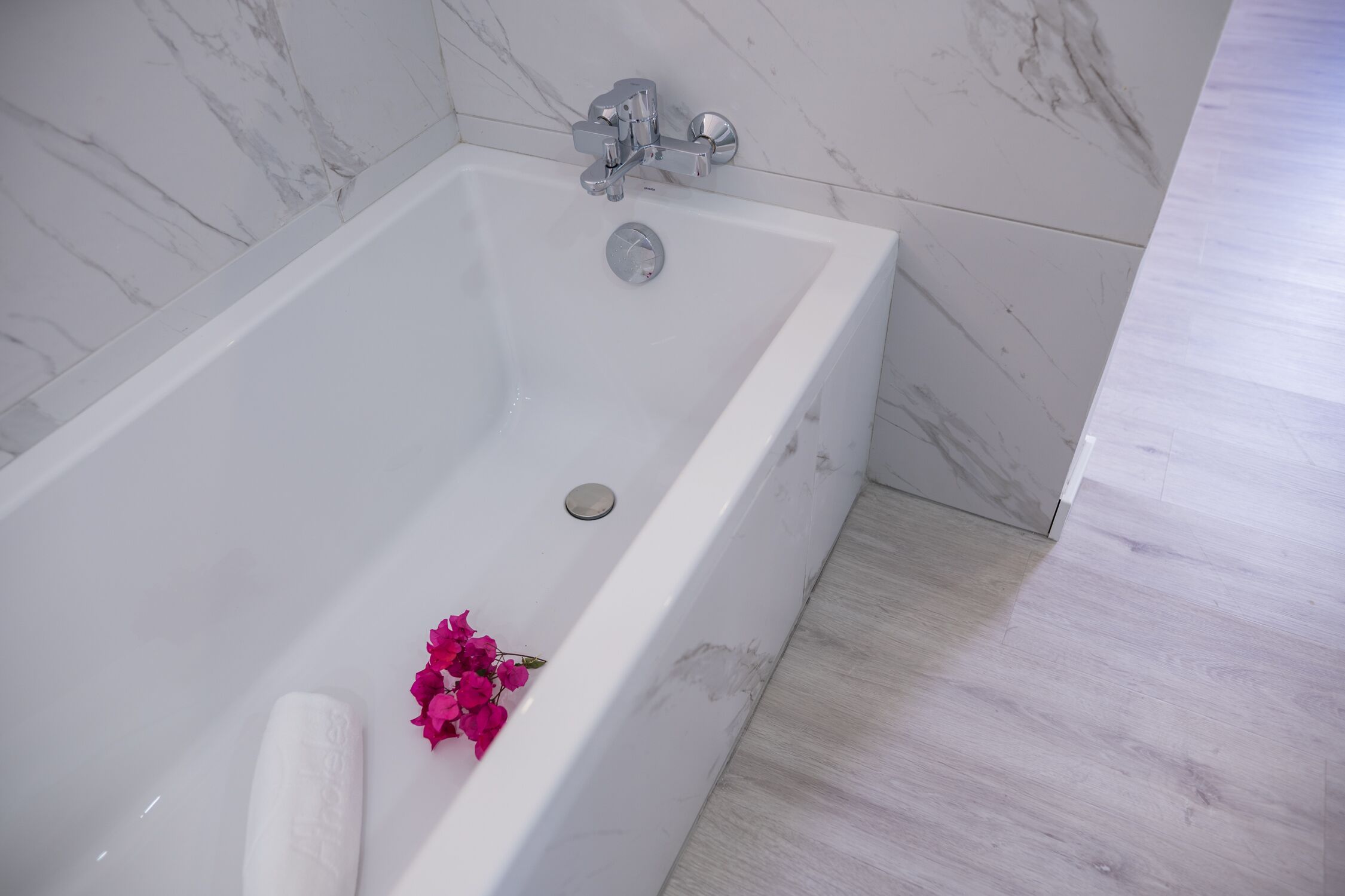 Una bañera blanca con una sola flor y una toalla enrollada, frente a una pared de mármol y suelo de madera.