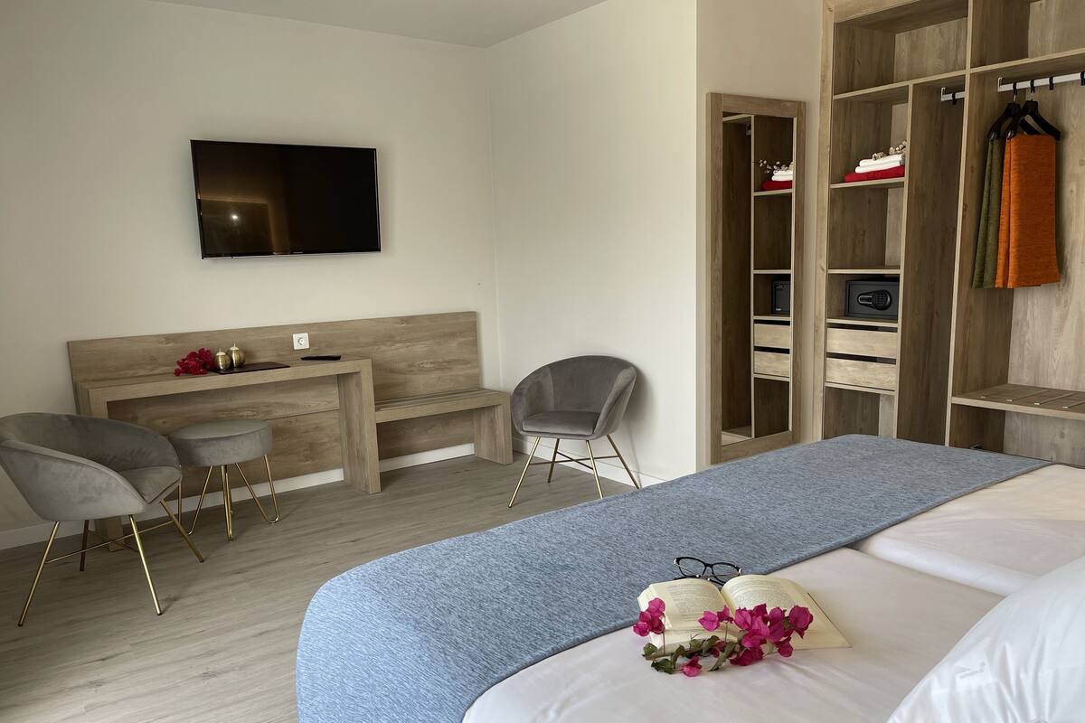A modern hotel room with a large bed, a desk with two chairs, a TV mounted on the wall, and a wooden wardrobe with hanging clothes.
