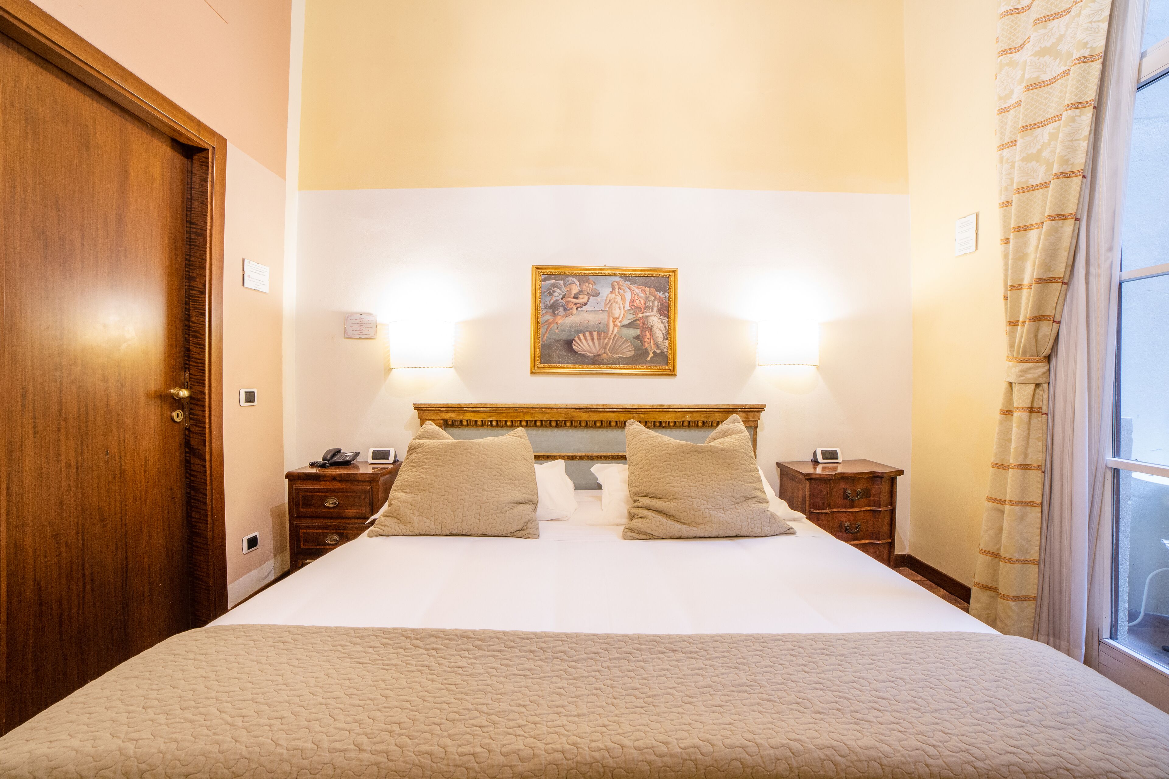 A hotel room with a large bed, two bedside tables, a nightstand, a wooden door, a painting on the wall, and a window with curtains.