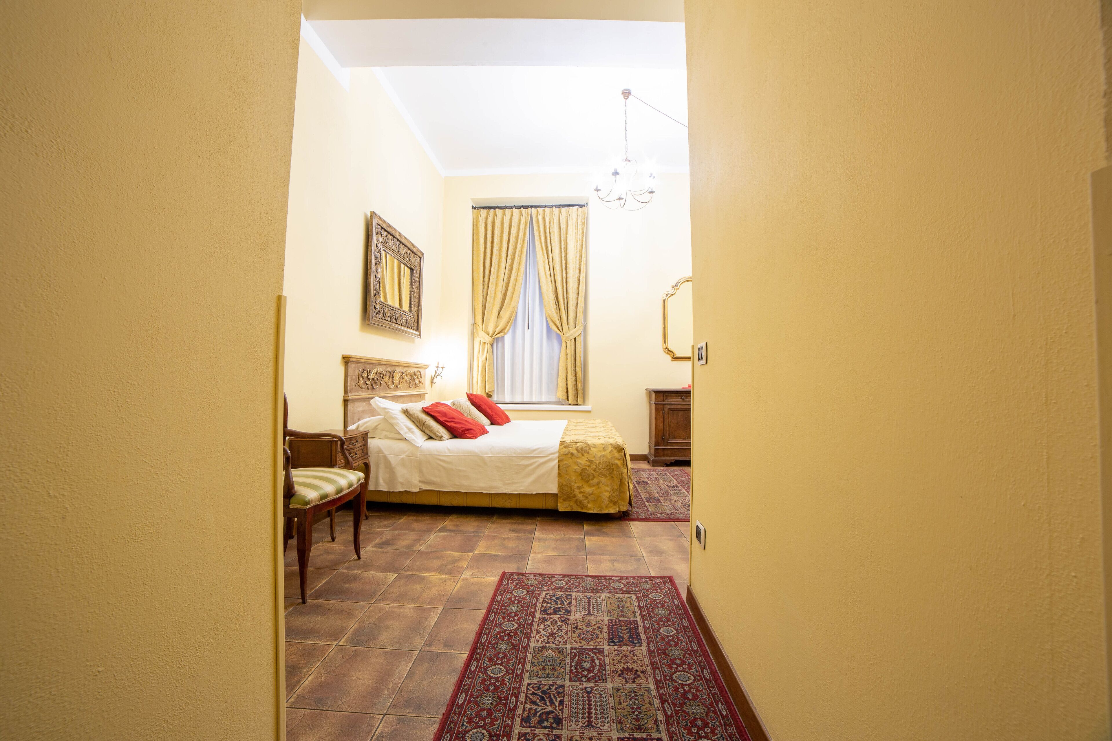 A bedroom with a bed, a chair, a window with curtains, a chandelier, and a patterned rug.
