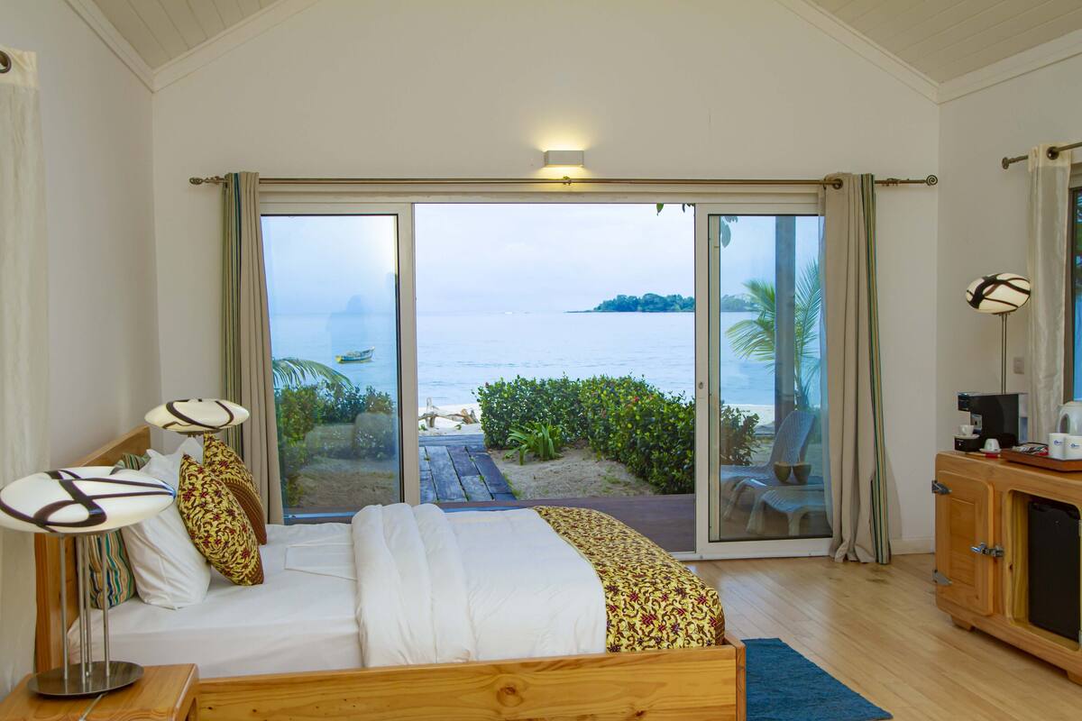 Ein Schlafzimmer mit einem Bett, Nachttischlampen, einem Holzschrank und Blick auf den Strand durch große Schiebeglastüren.