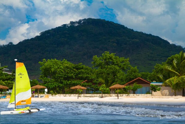 Una playa con un velero, palmeras y una montaña al fondo.