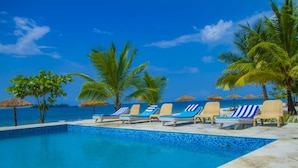 Un espace piscine agrémenté de chaises longues et de parasols, entouré de palmiers et offrant une vue sur l’océan.