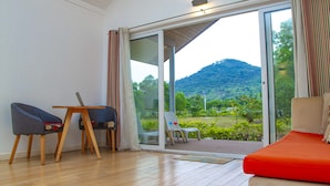 Une pièce avec un plancher en bois, un canapé rouge et une table entourée de chaises. Une porte-fenêtre coulissante donne sur un balcon d’où l’on peut admirer un paysage verdoyant et des collines.