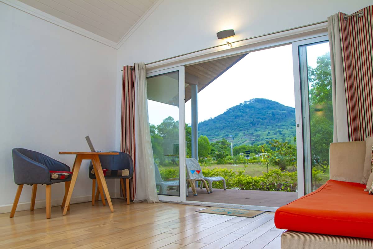 Ein Zimmer mit Holzboden, einem roten Sofa und einem Tisch mit Stühlen. Eine Glastür führt auf einen Balkon mit Blick auf Grünflächen und Hügel.