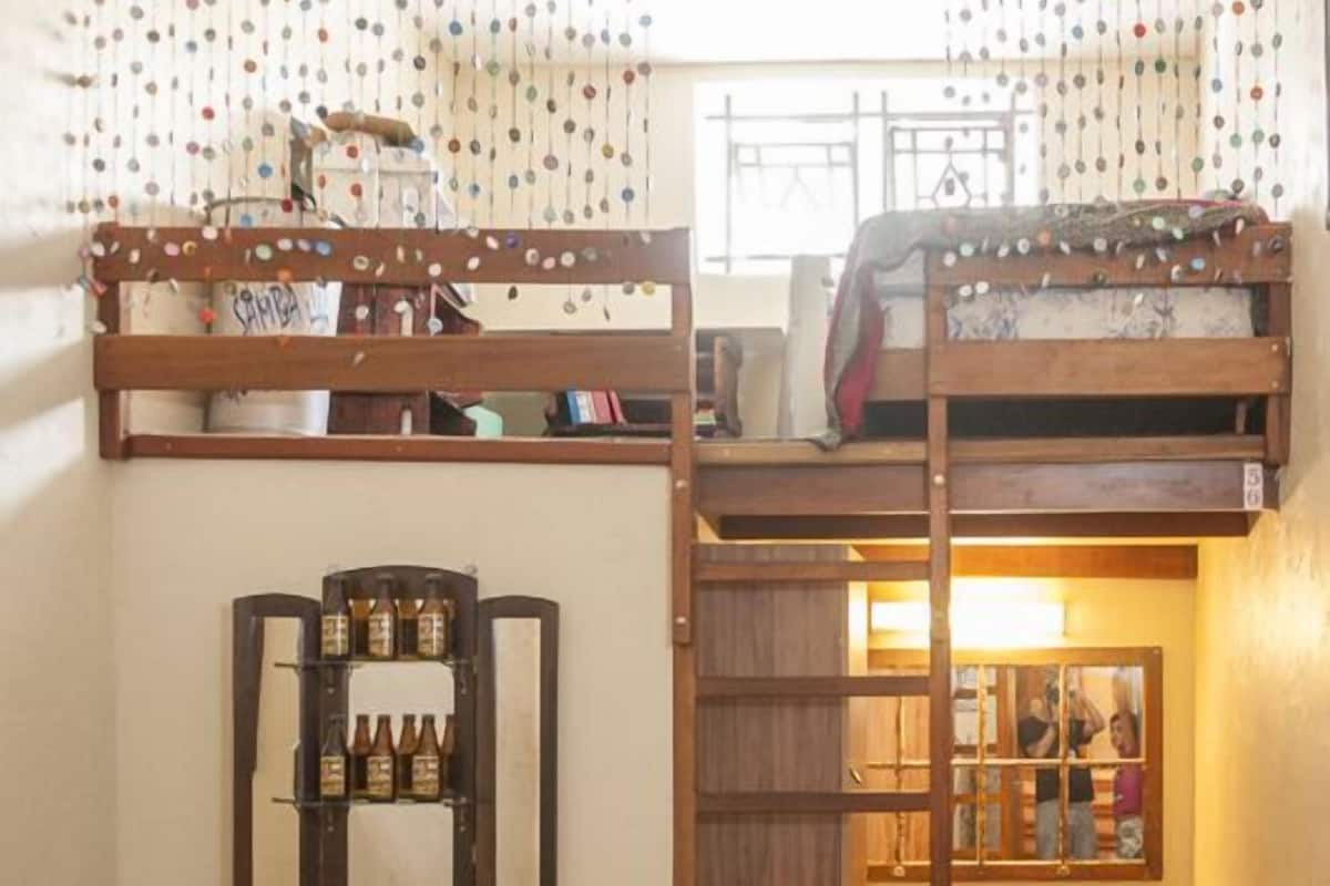 Une petite pièce comprenant un lit superposé, une échelle, une petite table et une étagère avec des bouteilles.