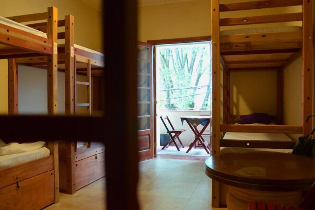 Une chambre avec un lit superposé en bois, une petite table et des chaises, une fenêtre donnant sur les arbres et une porte menant à l’extérieur.
