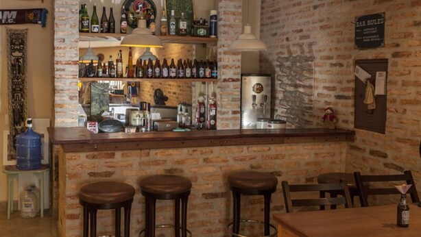 A bar area with a brick wall, shelves with bottles, a refrigerator, and stools. - Escambo Hostel (Sao Paulo)