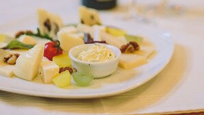 A plate of cheese and fruits on a table with a bottle and glasses in the background. - Mozart Hotel (Szeged)