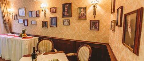 A dining area with a table set for two, a bottle of wine, and a plate of food. The room has patterned wallpaper, framed pictures on the walls, and a chandelier.