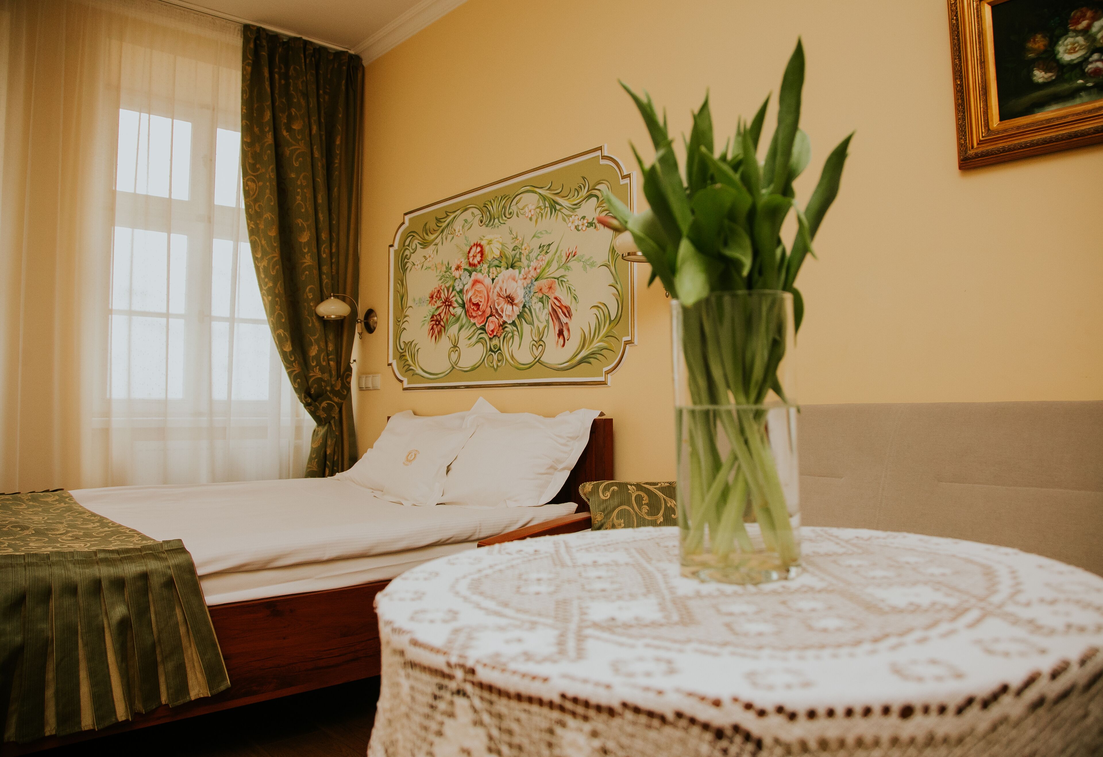 A bedroom with a bed, a bedside table with a vase of green plants, a wall with a floral painting, and a window with sheer curtains.