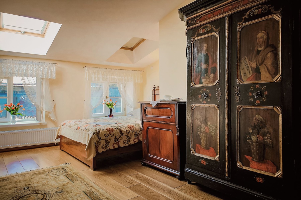 A room with a bed, a wooden cabinet with religious paintings, a window with lace curtains, and a skylight.