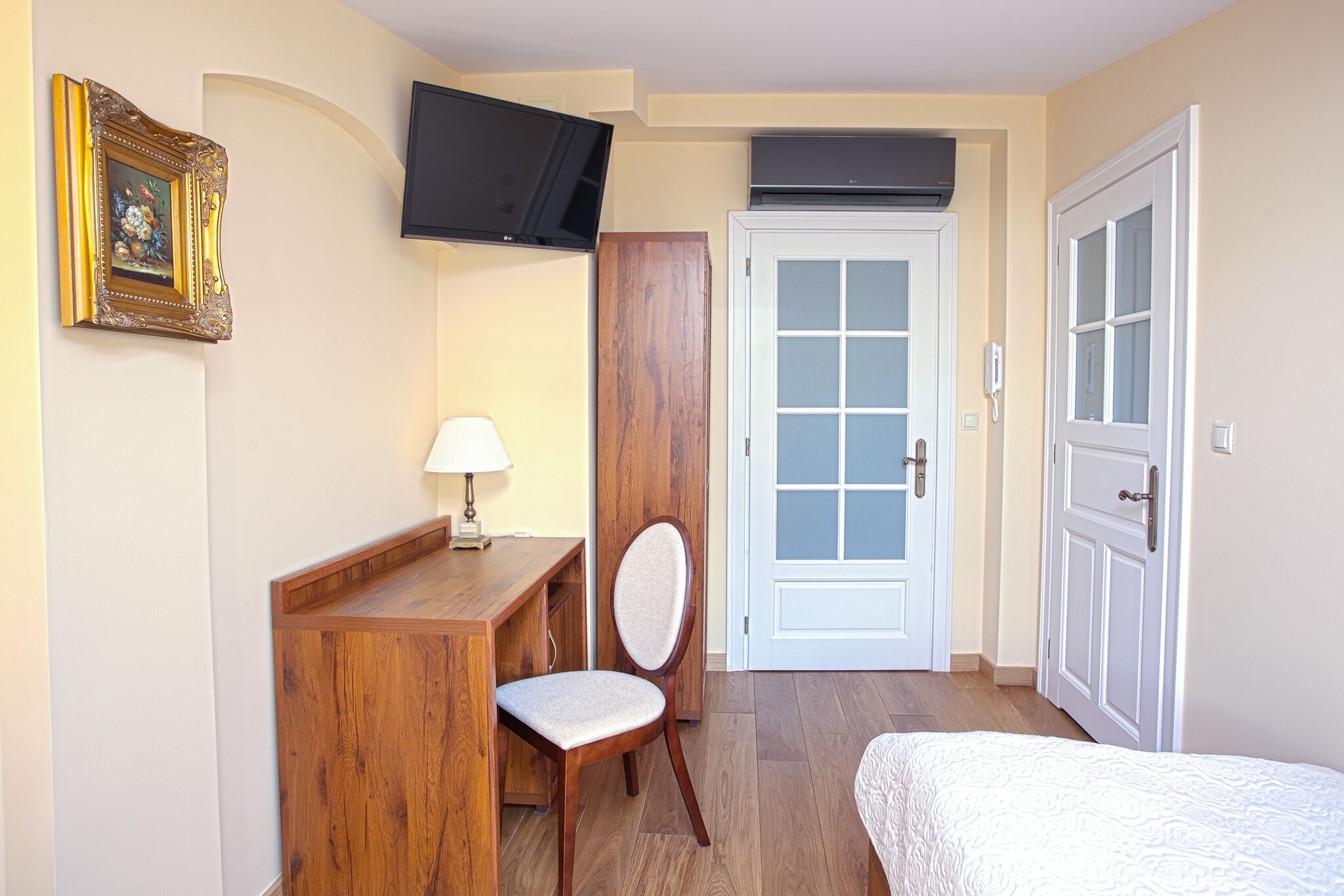 Una habitación con un escritorio de madera, una silla, una cama, un televisor montado en la pared y una puerta con paneles de vidrio esmerilado.