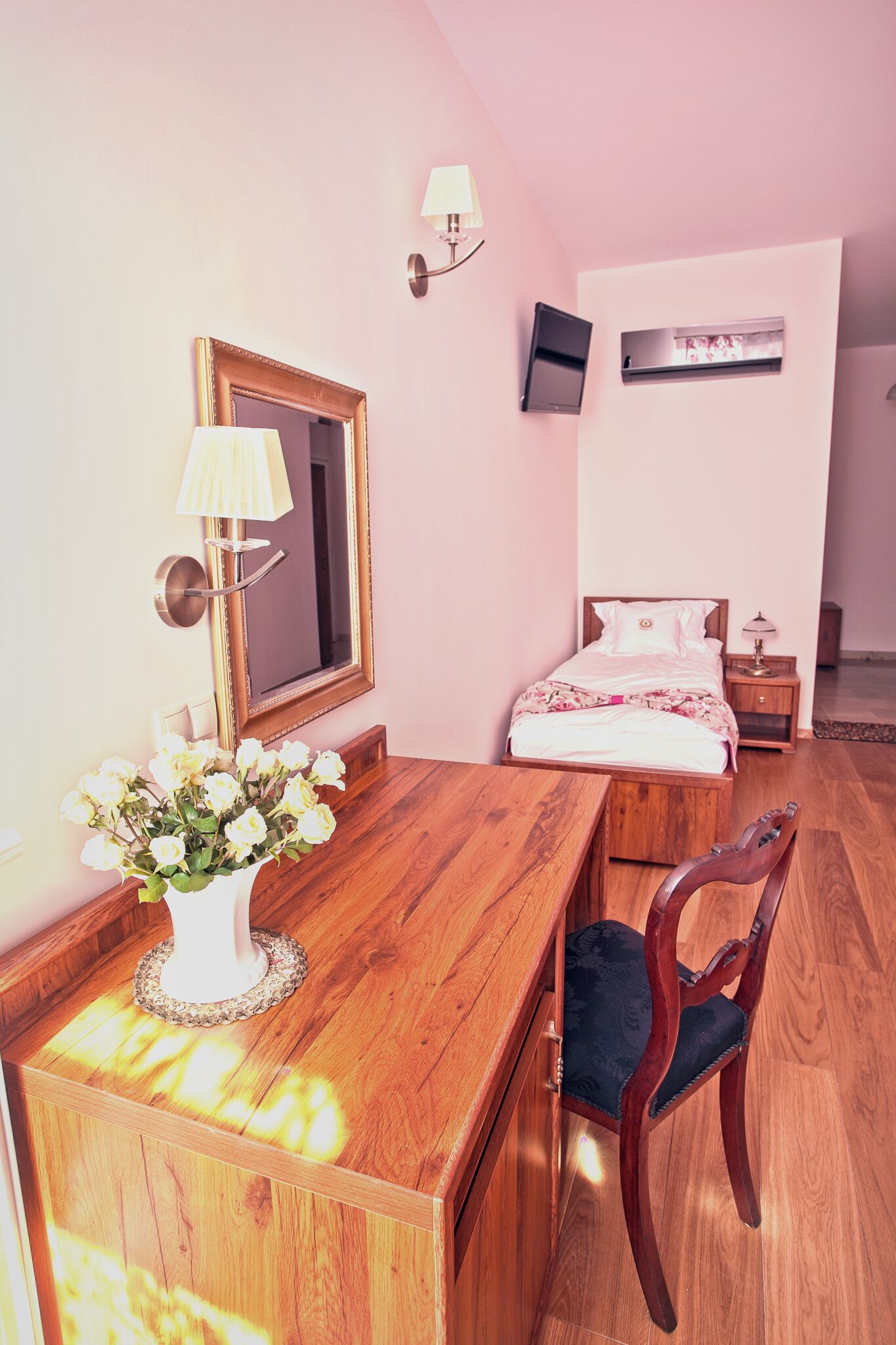 Une chambre avec un bureau en bois, une chaise, un lit recouvert d’une courtepointe blanche, une table de chevet avec une lampe, un téléviseur fixé au mur et un miroir.