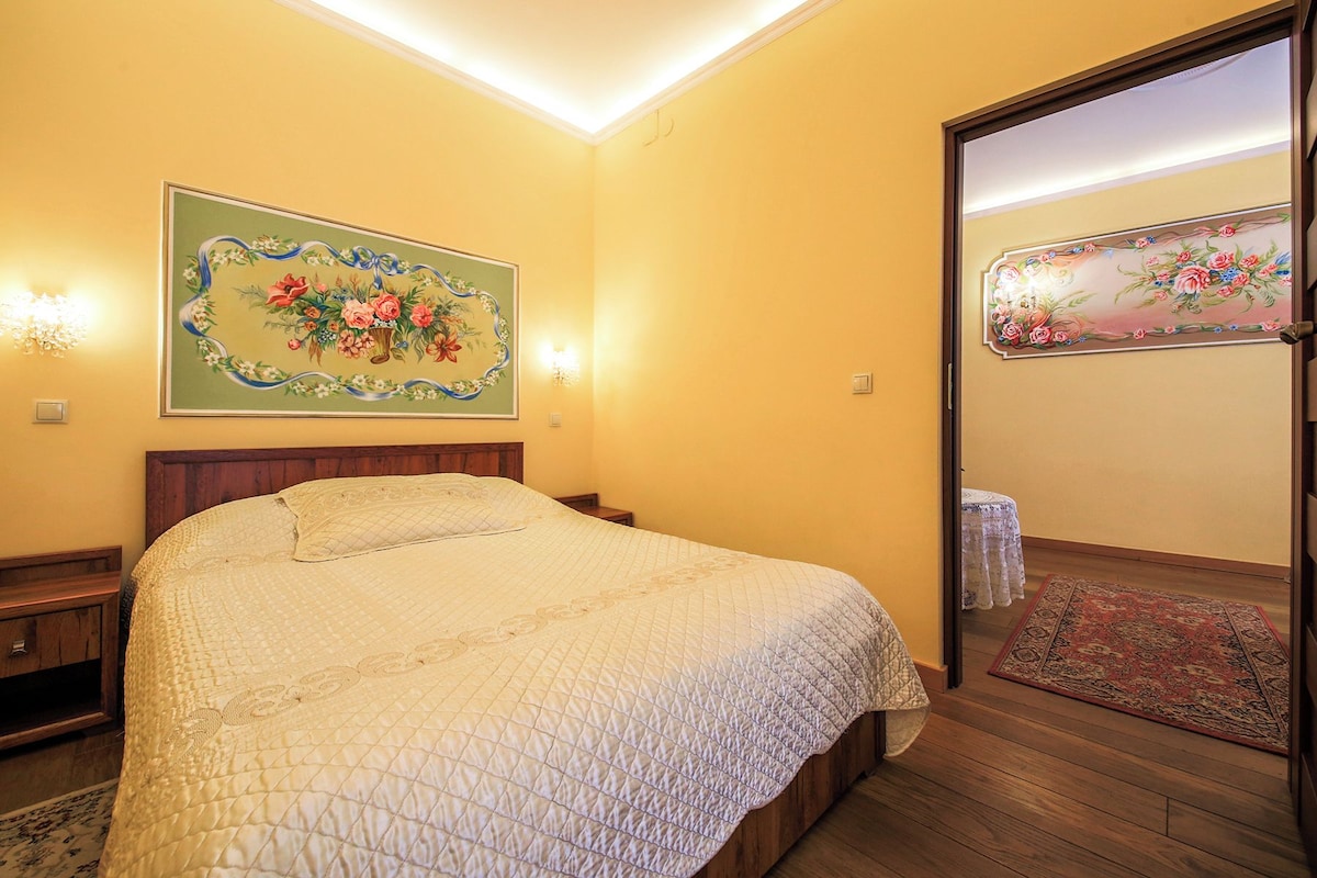 Un dormitorio con una cama, cabecera de madera, mesita de noche, lámpara de pared, un cuadro floral y una puerta con espejo.