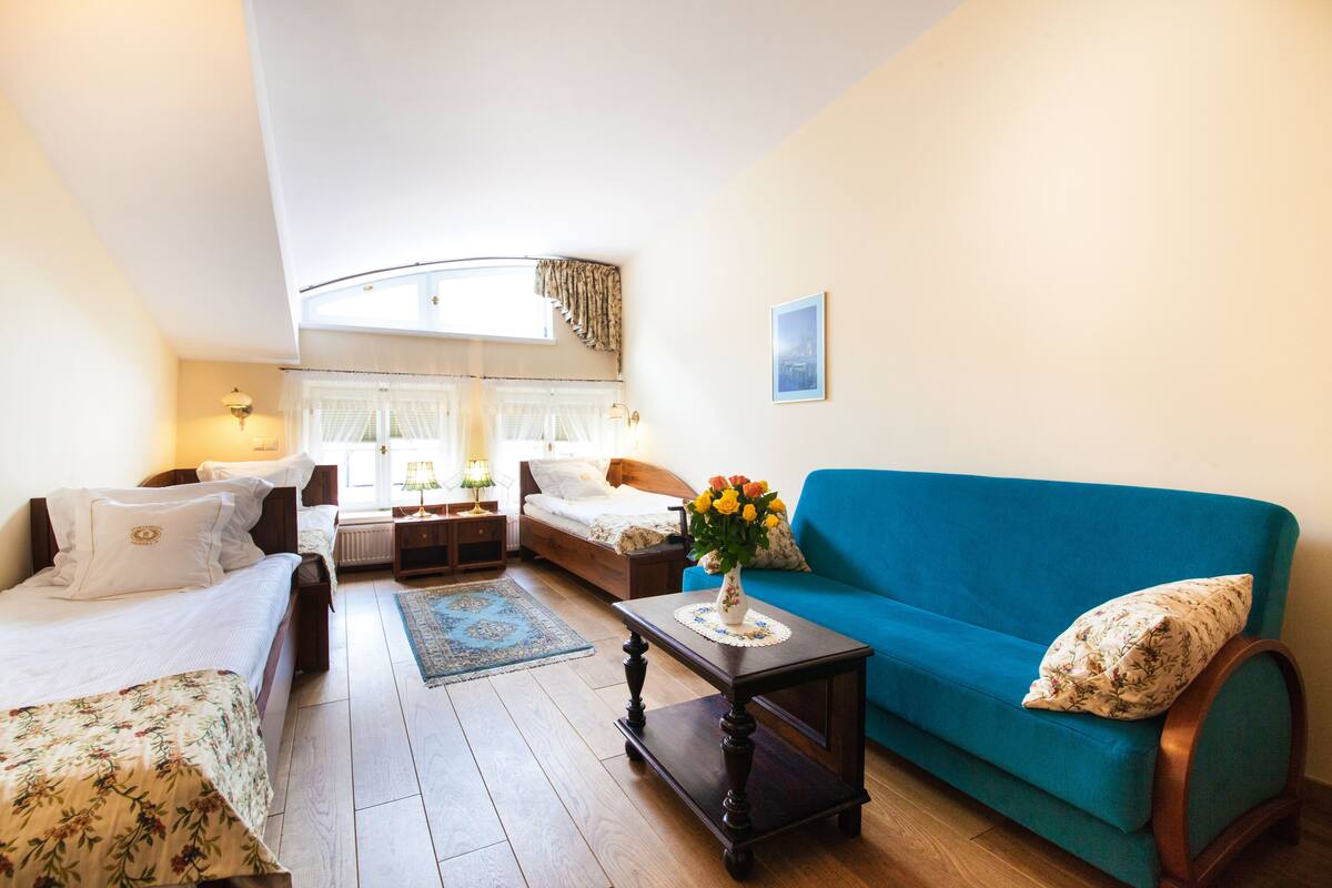 Een gezellige kamer met een bed, een blauwe bank, een kleine tafel met een bloemenvaas en een raam met gordijnen.