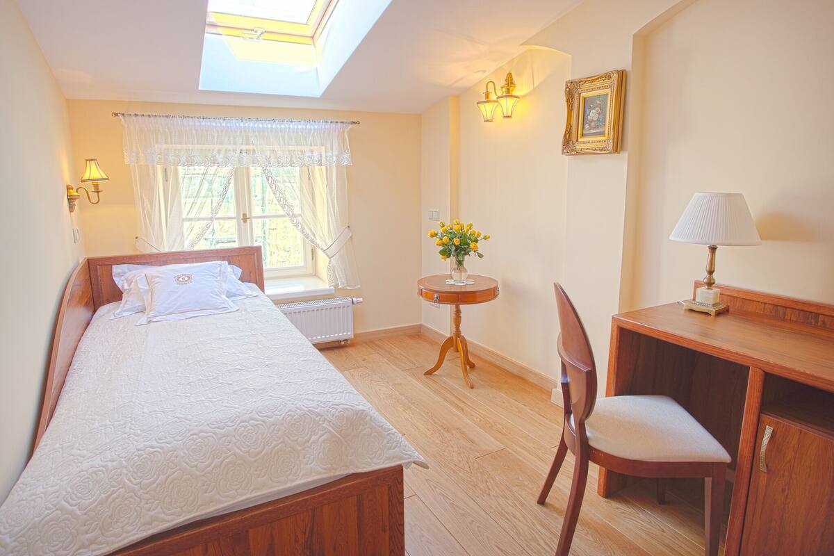 Een slaapkamer met een bed, een bureau, een stoel, een kleine tafel met bloemen en een raam met gordijnen.