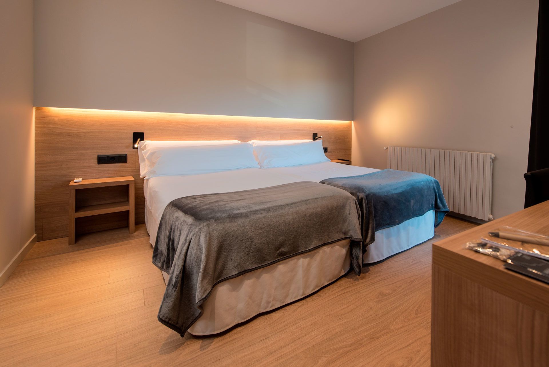 Una habitación de hotel moderna con cabecera de madera, una cama grande con dos almohadas, una mesita de noche y un aparador de madera.