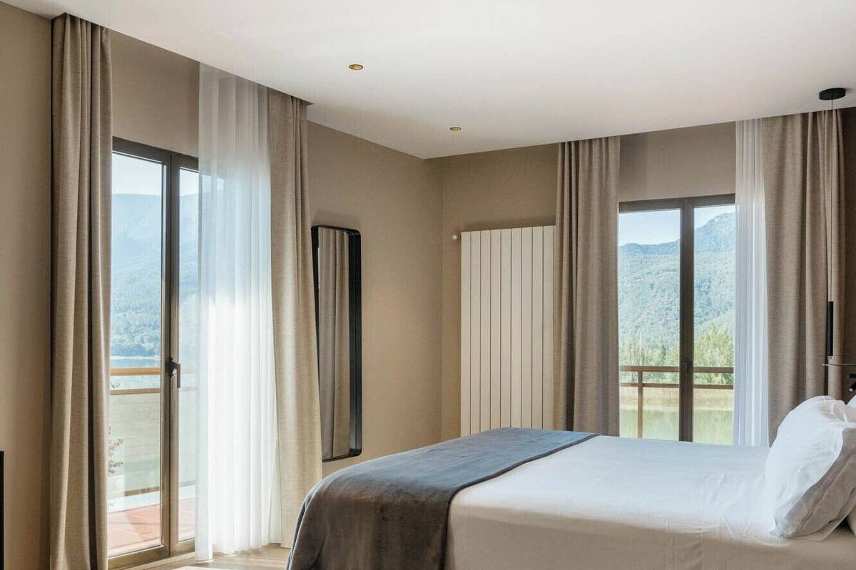 A modern bedroom with a large bed, a small side table, and a view of a mountainous landscape through the windows.