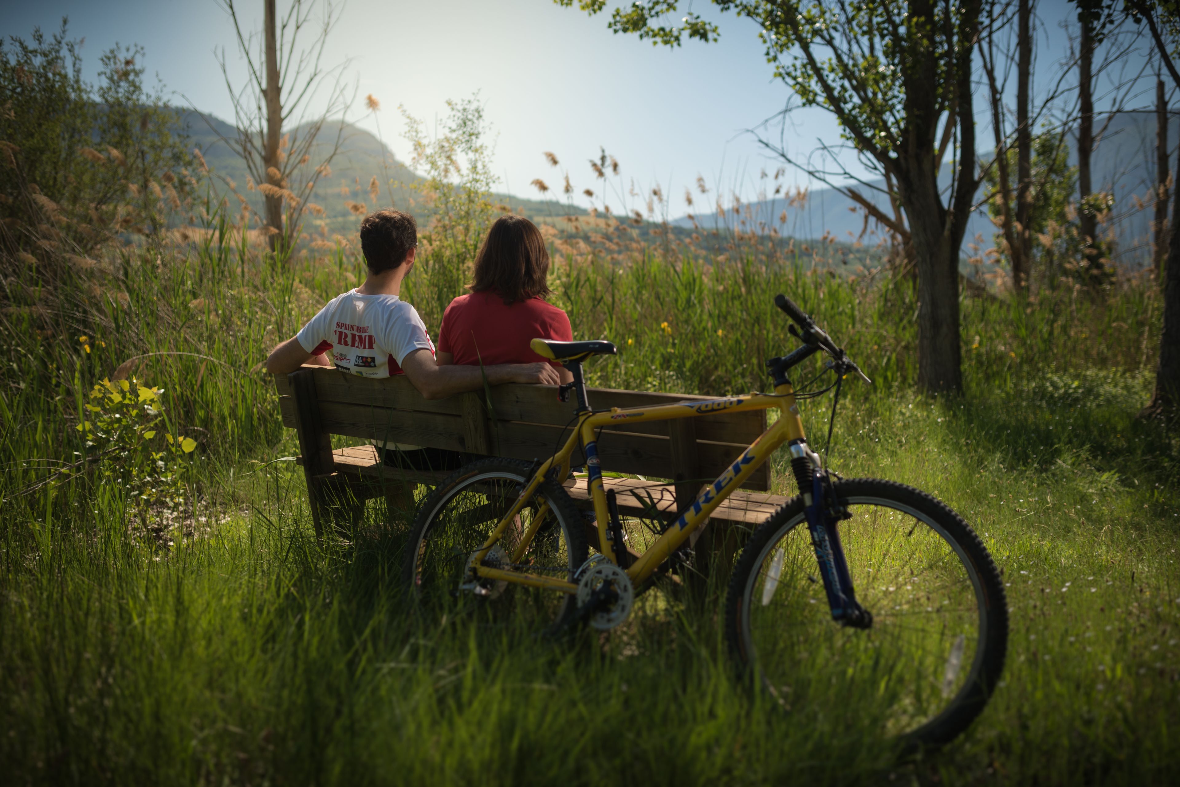 Two people sitting on a bench with a bicycle beside them in a grassy area with trees and mountains in the background.