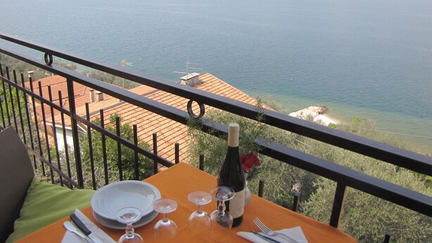 Una mesa con una botella de vino, copas y cubiertos, con vistas a la costa y montañas al fondo.