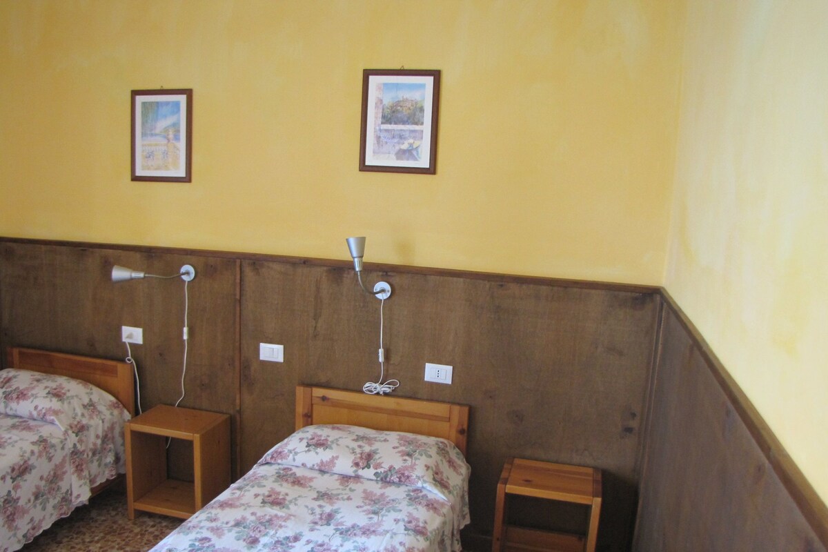 Una habitación con dos camas, cada una con cabecero de madera y una mesita de noche pequeña. Hay dos luces fijadas a la pared y dos cuadros enmarcados.
