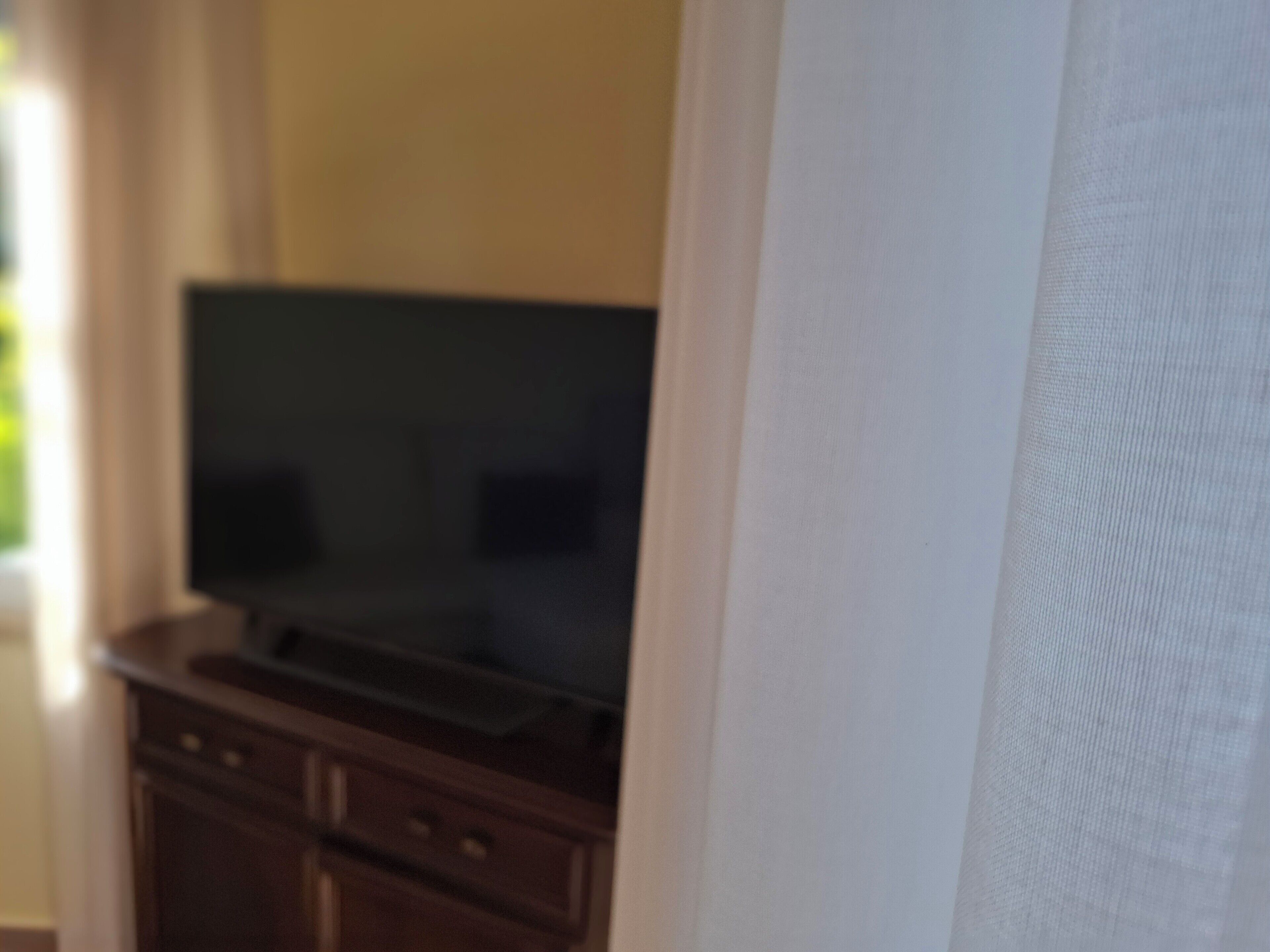 Ein Zimmer mit Fernseher auf einer Holzkassette, einem weißen Vorhang und einem Fenster mit Natürlichem Licht.