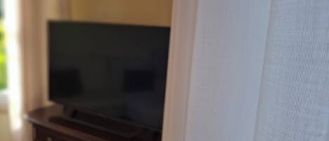 Ein Zimmer mit Fernseher auf einer Holzkassette, einem weißen Vorhang und einem Fenster mit Natürlichem Licht.