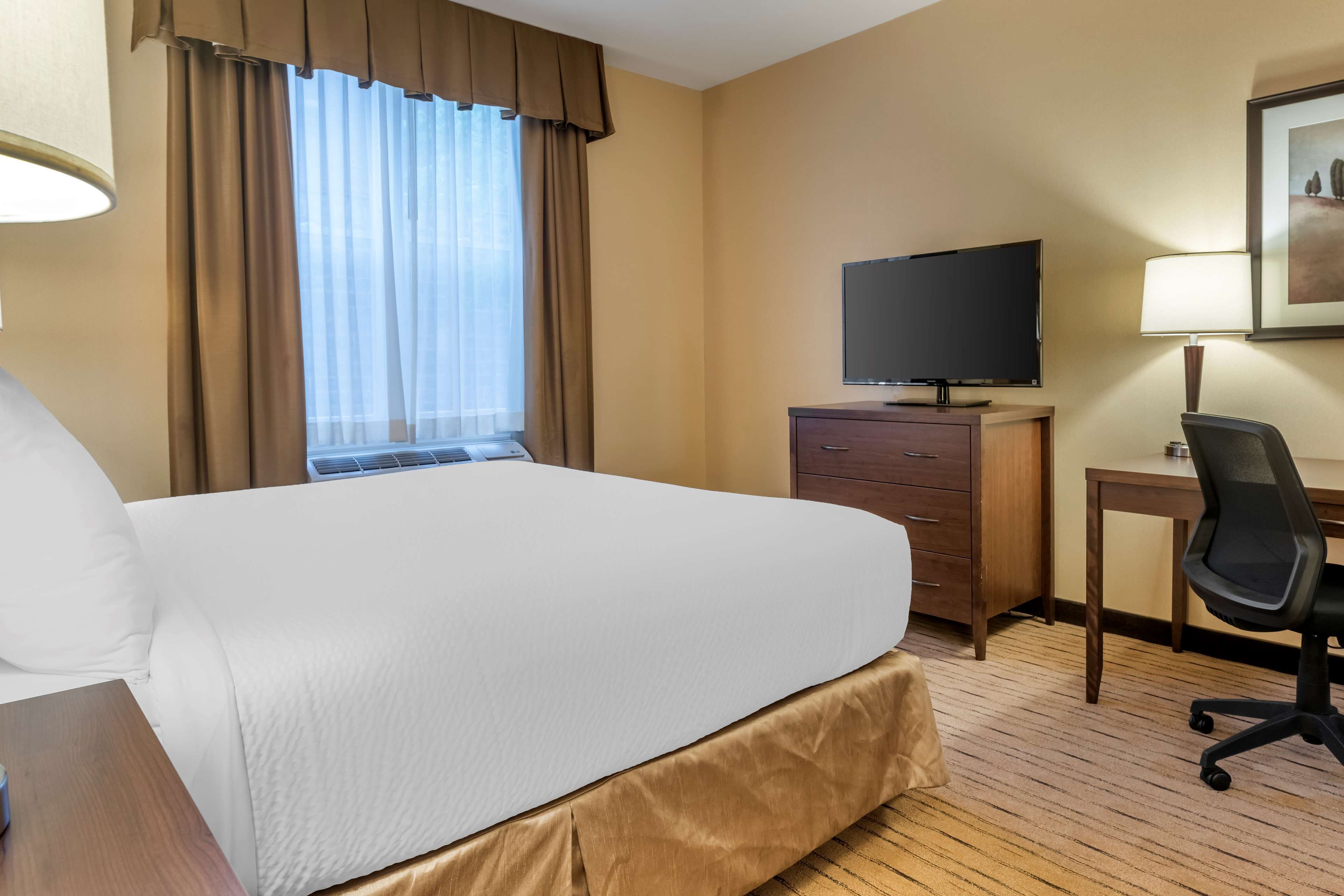 A hotel room with a large bed, a television on a wooden dresser, a desk with a chair, and a window with curtains.