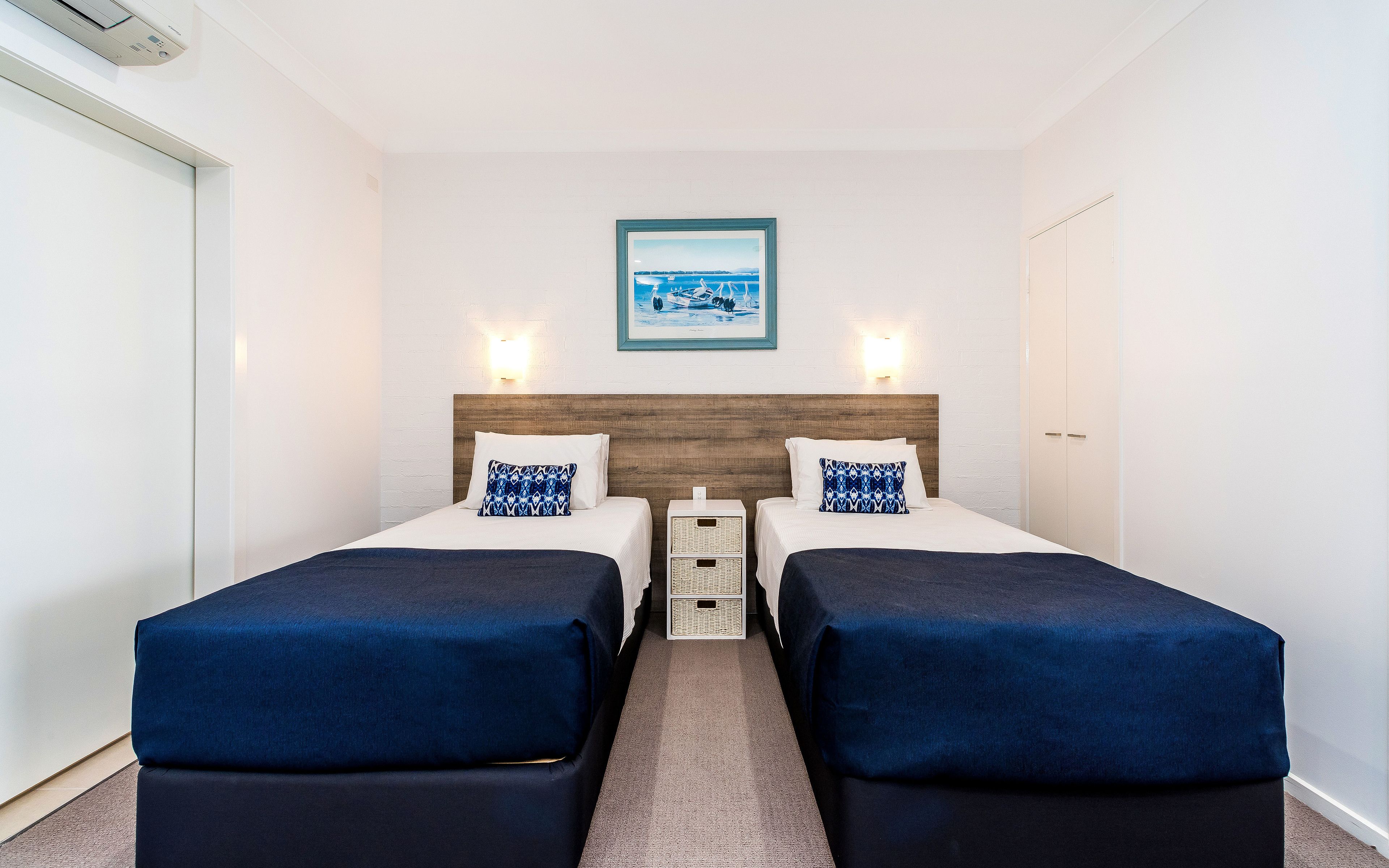 Quarto de hotel com duas camas, uma cabeceira de madeira, um quadro na parede e uma mesinha de cabeceira branca entre as camas.
