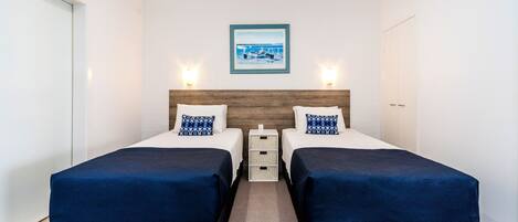 A hotel room with two beds, a wooden headboard, a framed picture on the wall, and a small white nightstand between the beds.