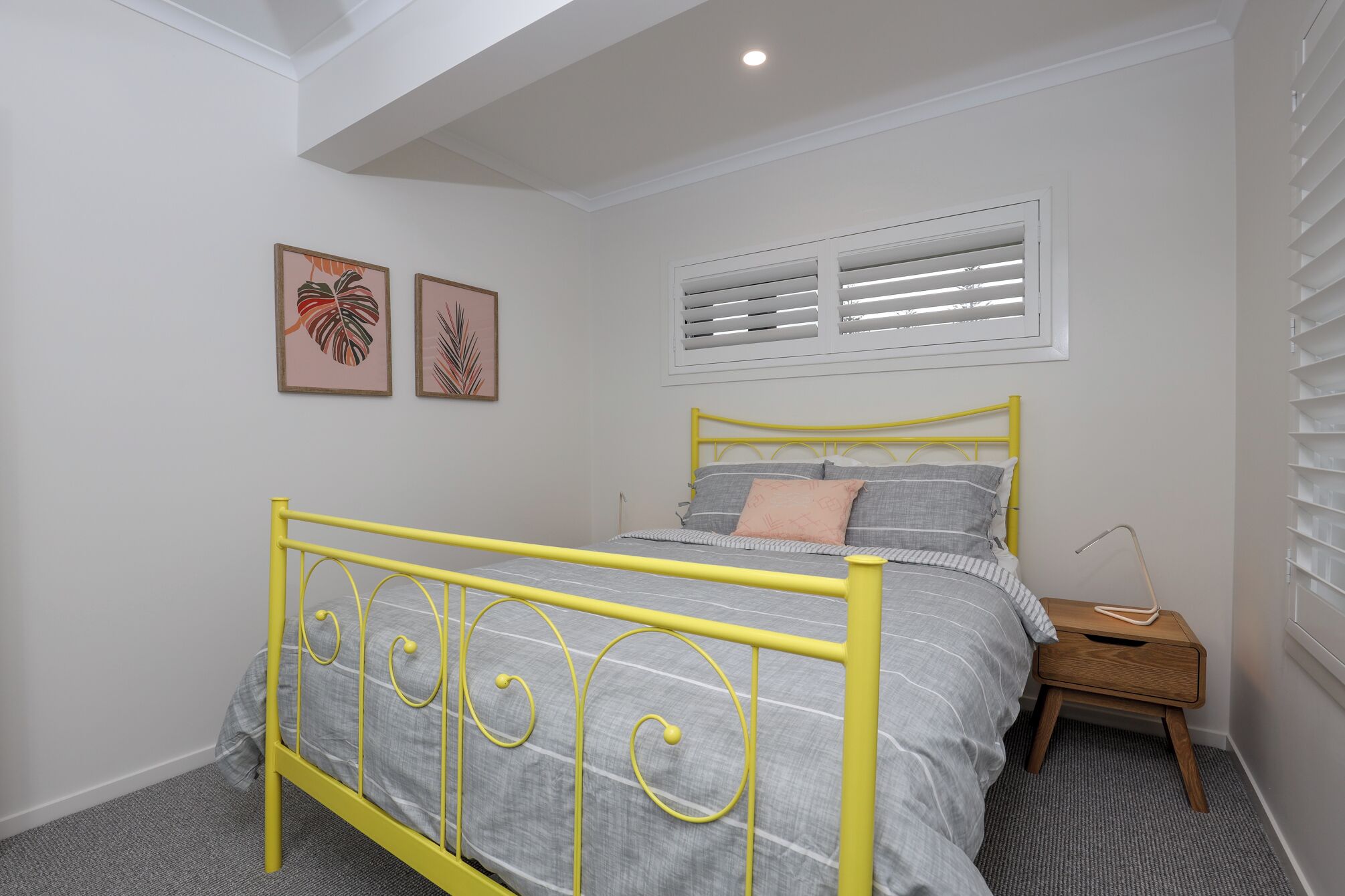 Een slaapkamer met een geel metalen bedframe, grijze beddengoed, twee kunstwerken met bladmotieven, een raam met zonwering en een nachtkastje met een lamp.
