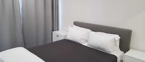 A neatly made bed with white bedding, a gray headboard, and a white nightstand with drawers.