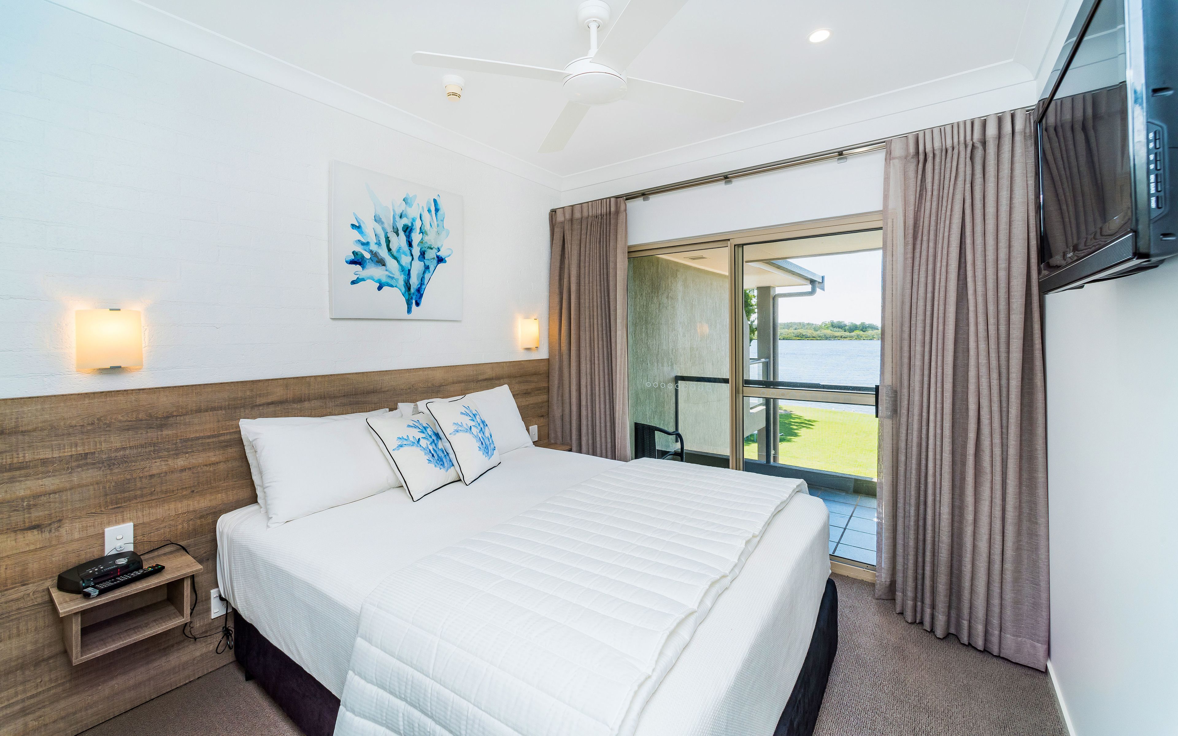 Quarto de hotel com cama, mesa de cabeceira, televisão na parede, ventoinha no teto e janela com cortinas.