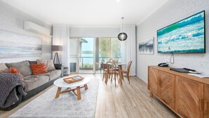 A modern living room with a sofa, coffee table, dining table, and chairs. There is a TV mounted on the wall, a wooden cabinet, and a large window with a view of the outdoors. - Moby Dick Waterfront Resort Motel (Yamba)