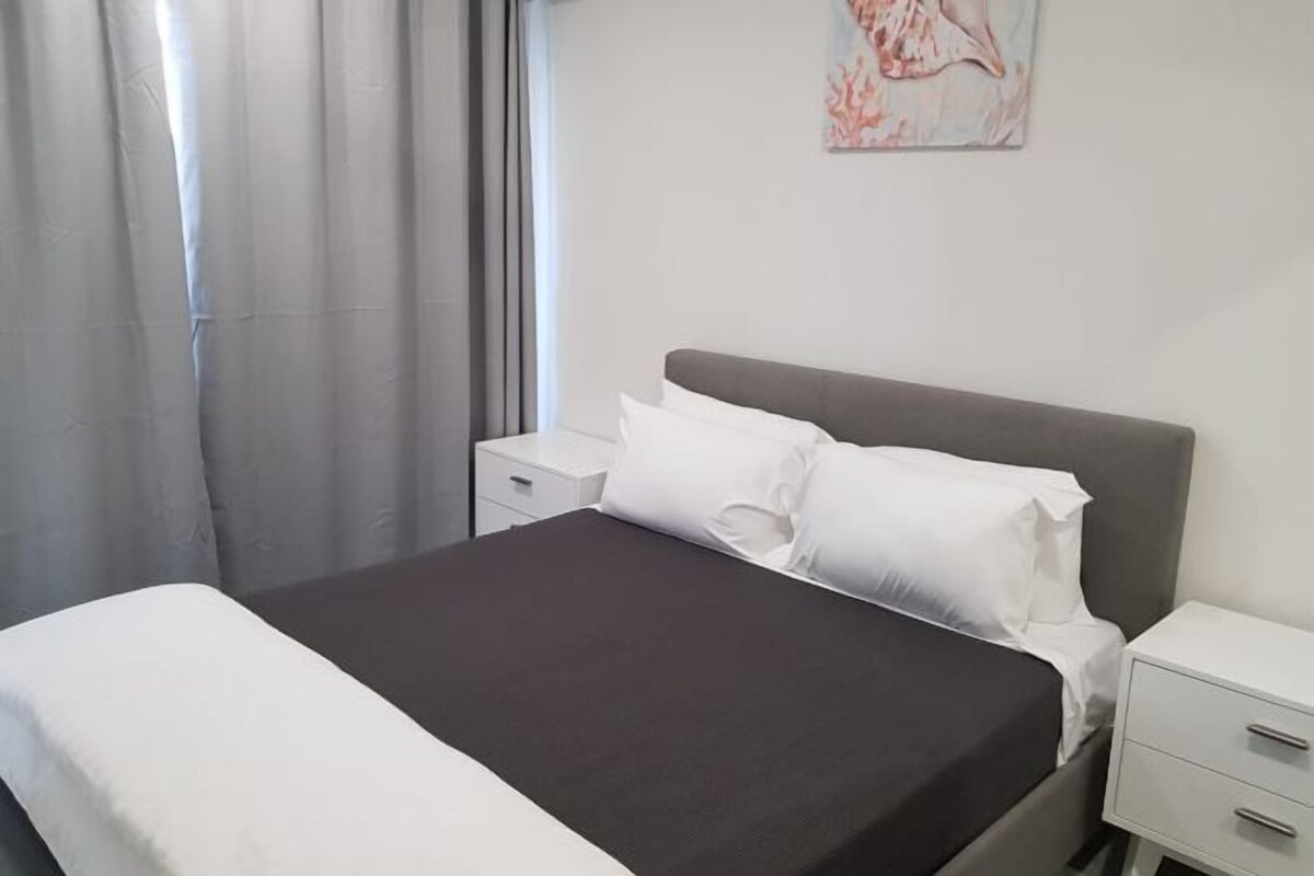 A neatly made bed with white and dark bedding, a bedside table with a drawer, and a wall-mounted artwork.