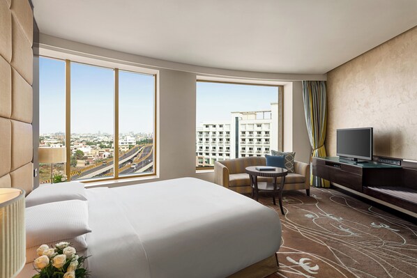 A hotel room with a large bed, a TV, a sofa, a small table, and a view of the cityscape. - Hilton Jaipur (Jaipur)