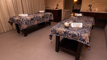 Two beds with 'AKASA LUXE' signs, a wooden bench, a mirror, and a stone wall.