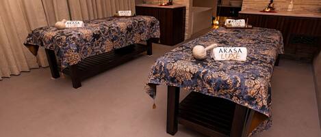 Dos camas con letreros de 'AKASA LUXE', un banco de madera, un espejo y una pared de piedra.