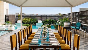 An outdoor dining area with a long table set for a meal, surrounded by chairs, with a pool and greenery in the background. - Hilton Jaipur (Jaipur)
