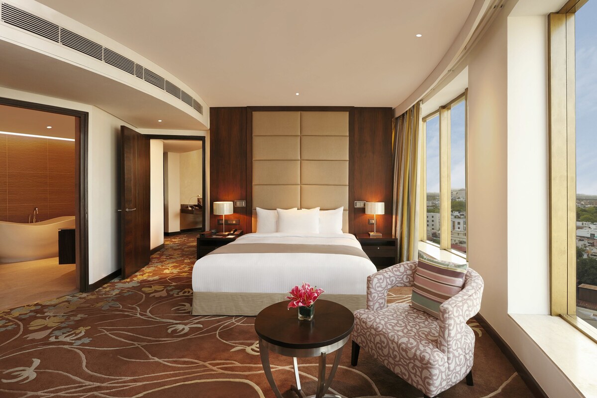 A hotel room with a large bed, a sitting area with a chair and table, a view of the city through large windows, and a bathroom with a bathtub and shower.