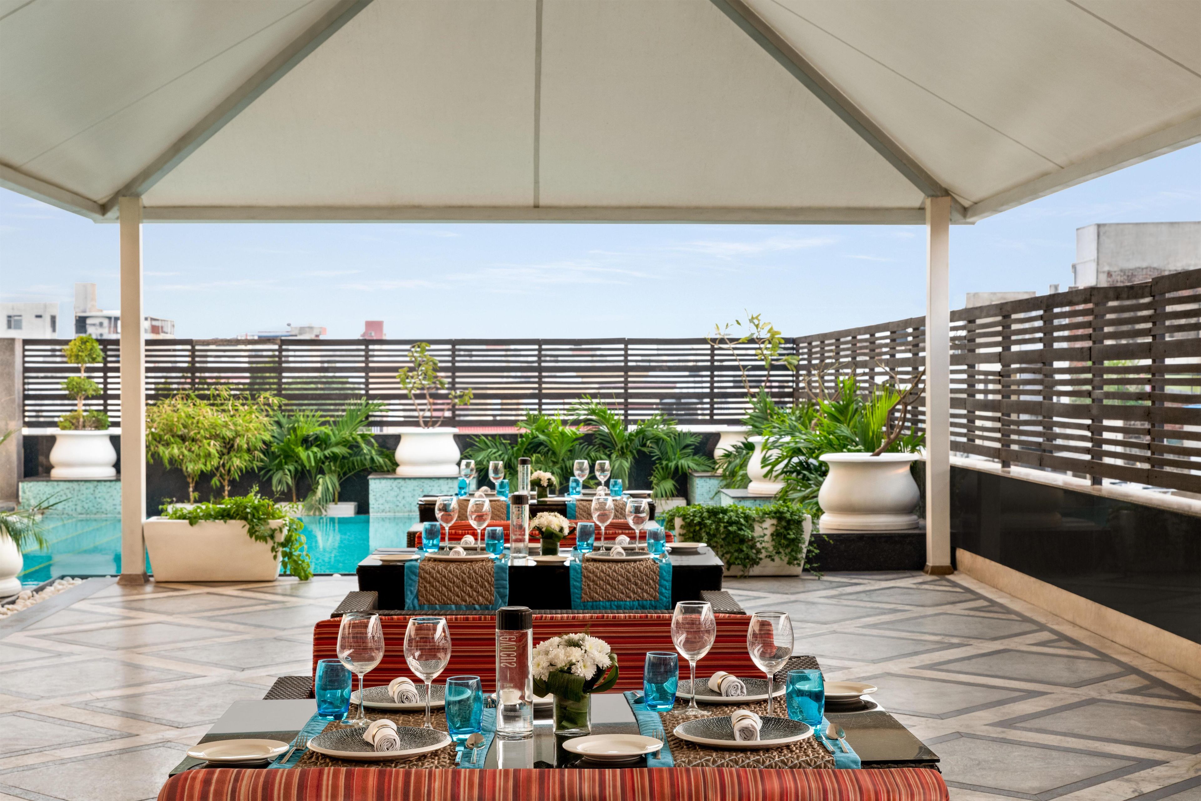 An outdoor dining area with tables set for two, surrounded by potted plants and a view of buildings.
