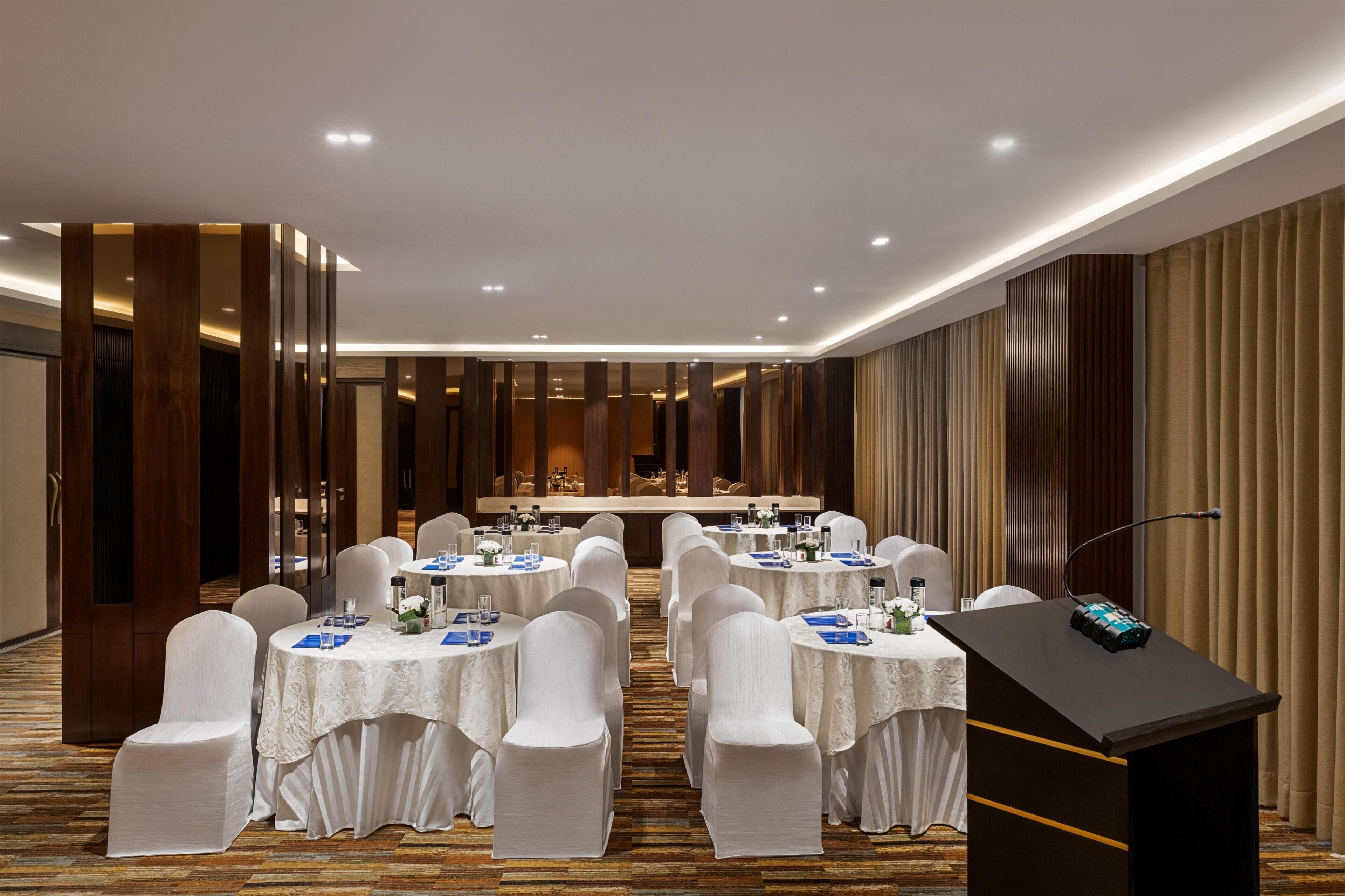 A conference room with round tables, white chairs, a podium, and a long table with a buffet setup.