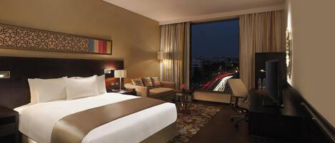 A hotel room with a large bed, a sofa, a desk with a TV, a lamp, and a view of the cityscape at night.