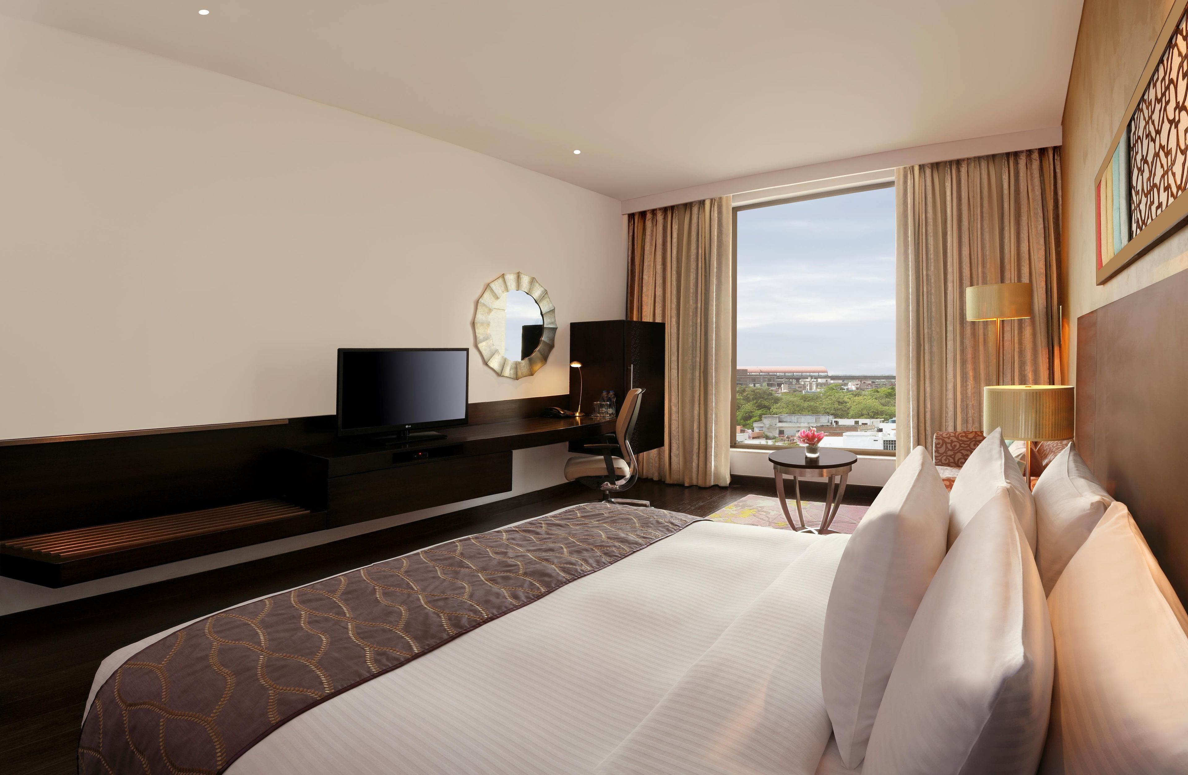 A hotel room with a large bed, a TV, a desk, a chair, a lamp, and a view of the city.